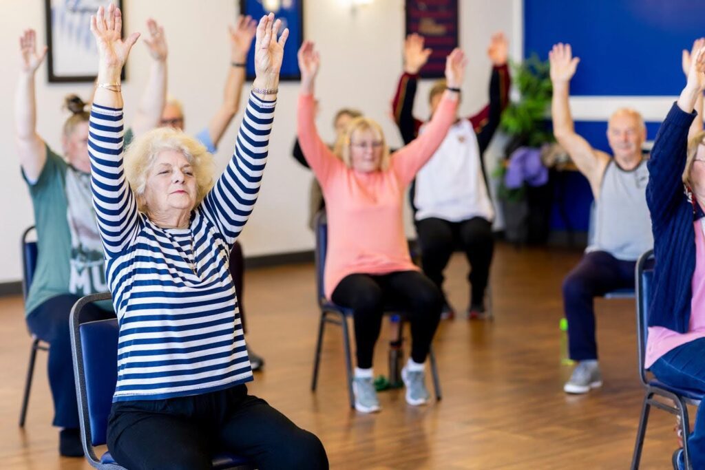 Group of seniors participating in a seated exercise class with arms raised, demonstrating ways to engage even sedentary older adults in physical activity.