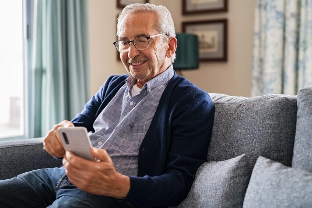 Smiling senior man sitting on a couch holding a smartphone, representing the use of reassuring phone script techniques to help older adults feel safe and supported.