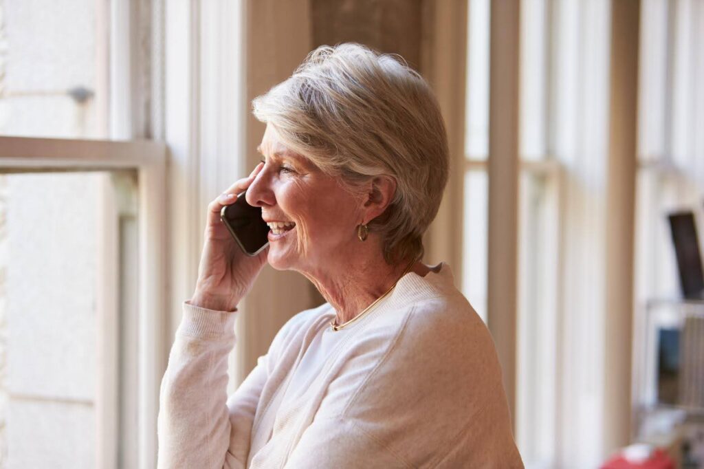 Smiling senior woman talking on the phone by a window, showing how some older adults feel more open sharing their thoughts over the phone than in person.