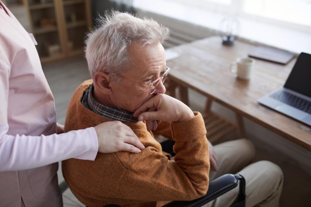 An elderly man in a wheelchair, looking thoughtful, with a caregiver offering comfort by placing a hand on his shoulder, symbolizing the emotional labor of aging.