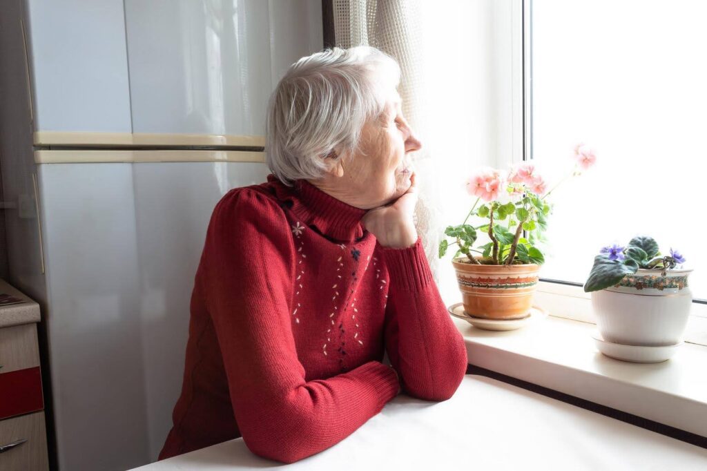 An introverted senior woman in a red sweater sitting by the window, quietly reflecting, showing the importance of gentle companion care for introverts.