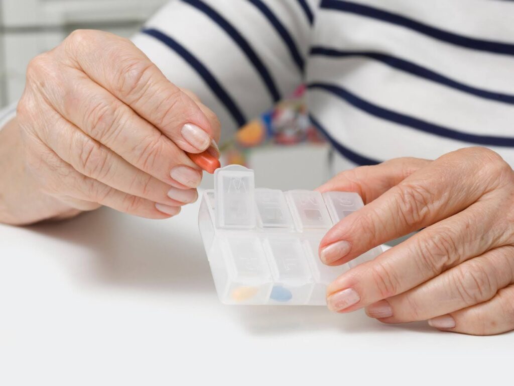 Senior organizing daily pills into a pillbox, comparing pillboxes vs human reminders for reliable medication management.
