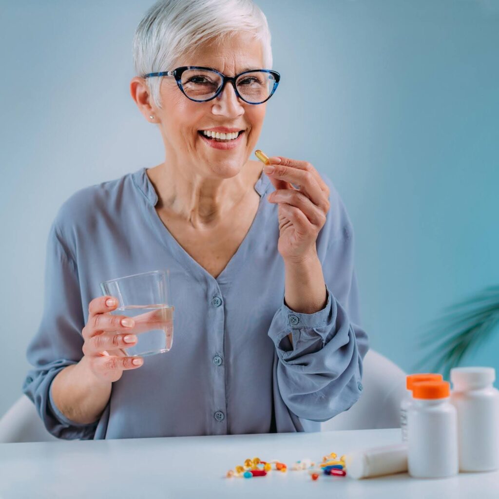 Smiling senior woman holding a pill and a glass of water with supplement bottles on the table, illustrating the discussion around supplements and senior safety.
