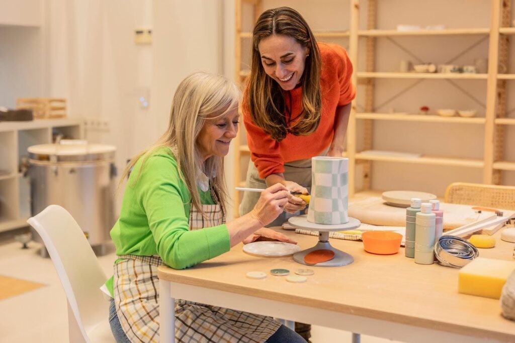 Senior woman enjoying creative activities with a caregiver, painting pottery together during a companion care visit.