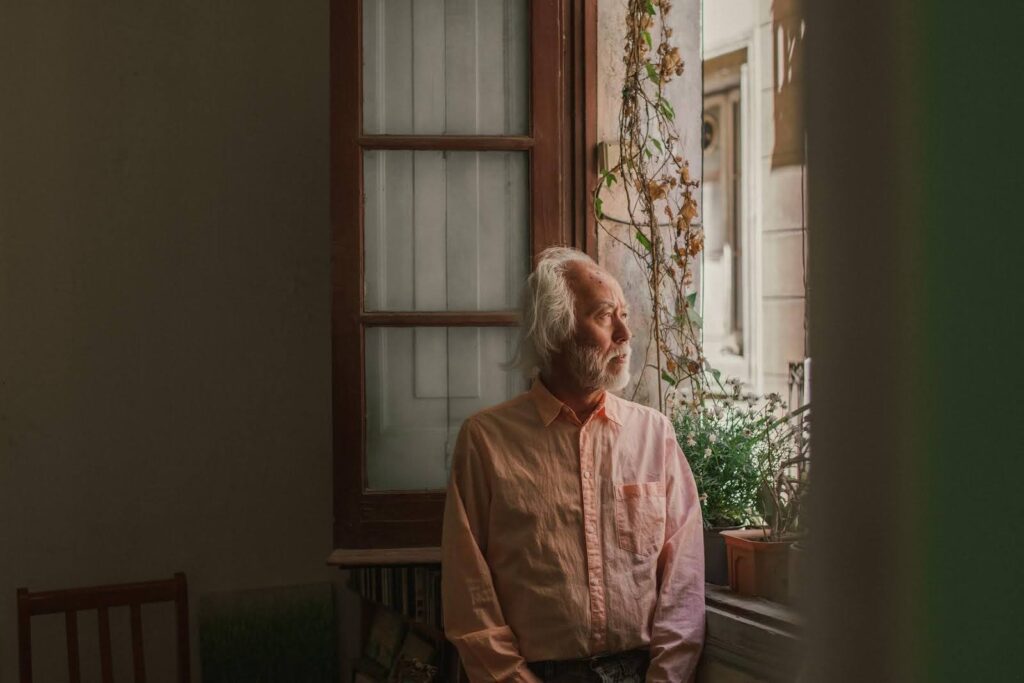 Elderly man standing by a window, looking outside thoughtfully, symbolizing grief, loneliness, and the importance of emotional and mental wellness support for seniors at home.