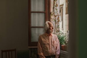 Elderly man standing by a window, looking outside thoughtfully, symbolizing grief, loneliness, and the importance of emotional and mental wellness support for seniors at home.