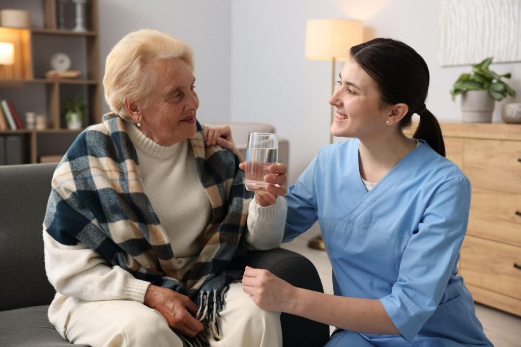 Caregiver in blue uniform offering a glass of water to a smiling elderly woman, illustrating the difference between home care and home health services.”