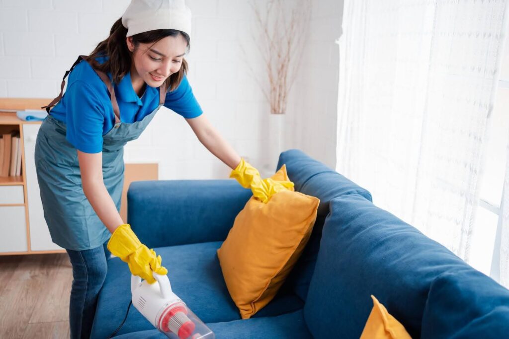 Smiling caregiver vacuuming a sofa and adjusting pillows, representing cleaning support for seniors at home.