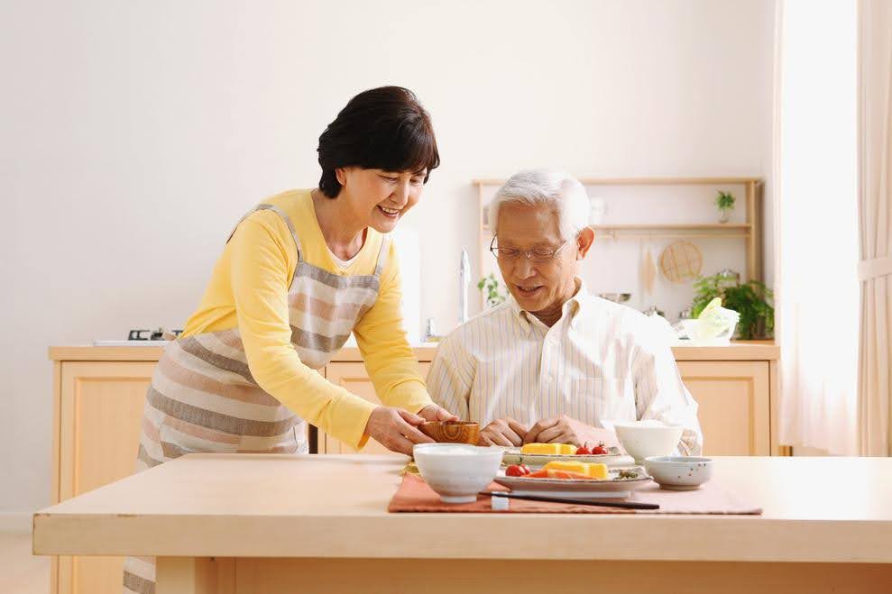 Smiling caregiver serving a healthy meal to an elderly man at a dining table, representing personalized meal planning and support for seniors with dietary restrictions.