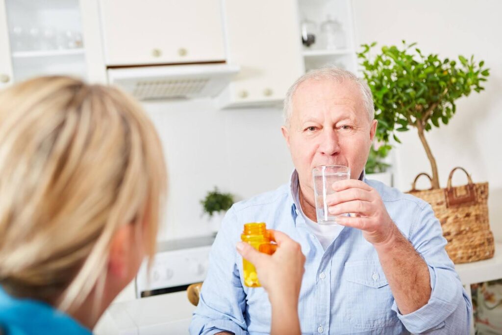 Senior man drinking water while caregiver offers medication, highlighting the challenge when parents refuse to take their prescribed meds.
