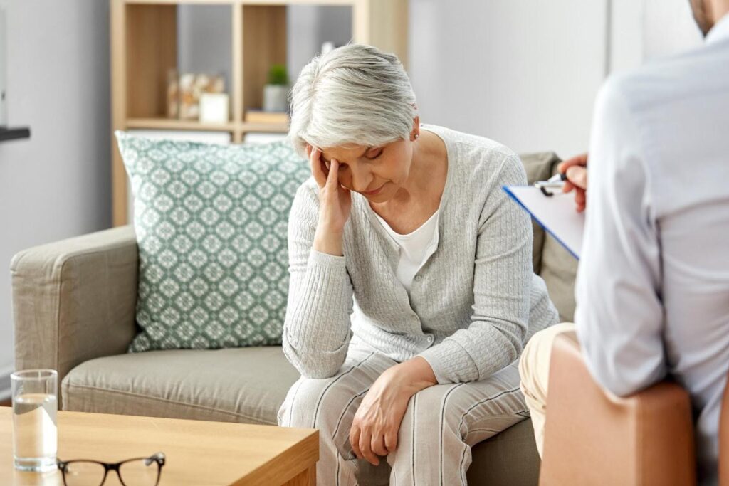 lderly woman sitting on a couch with her head in her hand while talking to a caregiver, symbolizing the difficulty of asking for help.