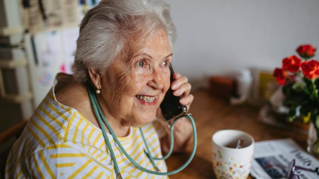 Smiling senior woman talking on the phone at home, symbolizing the positive psychological impact of regular check-ins and emotional connection for older adults.