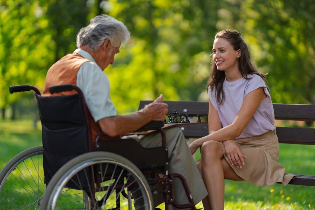 Young woman talking with an elderly man in a wheelchair at the park, addressing senior loneliness through companionship and empathy.