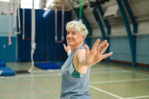 Smiling senior woman stretching with arms open in a gym, representing active aging and the fun, energetic approach of Zumba compared to traditional senior fitness routines.