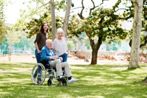 Family supporting an aging parent in a wheelchair while spending time together outdoors for Ace Home Care.