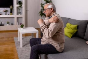 Elderly man experiencing shoulder discomfort while sitting at home, highlighting how Ace Home Care supports seniors with chronic illness and limited mobility in managing daily activities.
