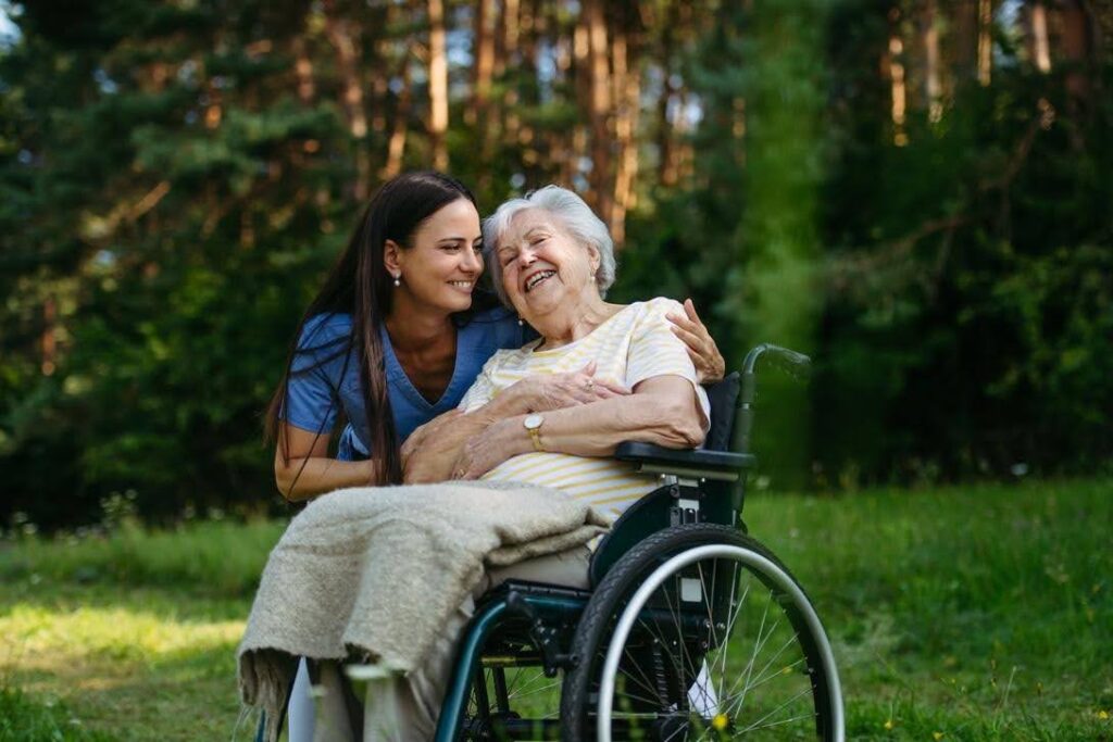 Caregiver for a senior with limited mobility providing companionship and emotional support to an elderly woman in a wheelchair outdoors.