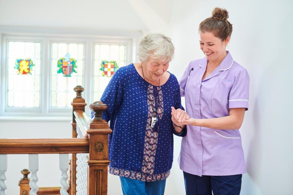 Caregiver assisting an aging parent with walking safely near the stairs at home, highlighting the need for additional senior care support beyond weekly family visits.